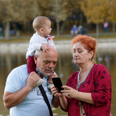 Na zdjęciu babcia i dziadek, przeglądający coś w smartfonie. Seniorzy opiekują się wnukiem.