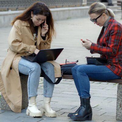 dwie kobiety siedzące na ławce, jedna korzysta z laptopa, a druga trzyma w ręce smartfon