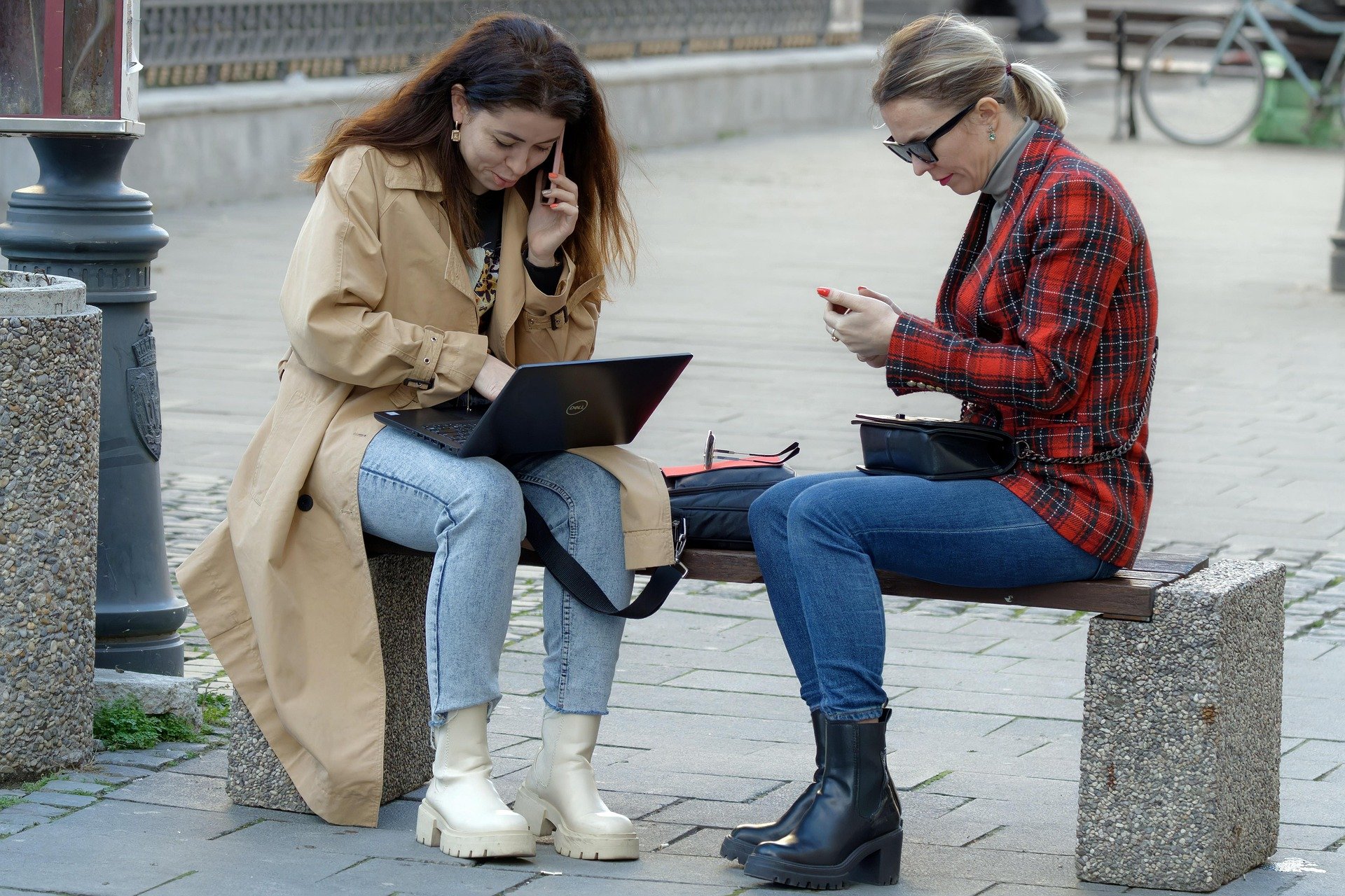 dwie kobiety siedzące na ławce, jedna korzysta z laptopa, a druga trzyma w ręce smartfon