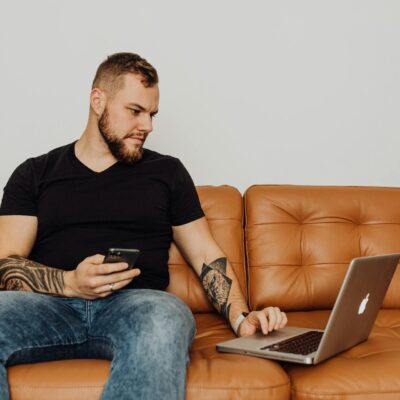 mężczyzna ze smartfonem w ręce siedzący na kanapie i drugą ręką robiący coś na laptopie